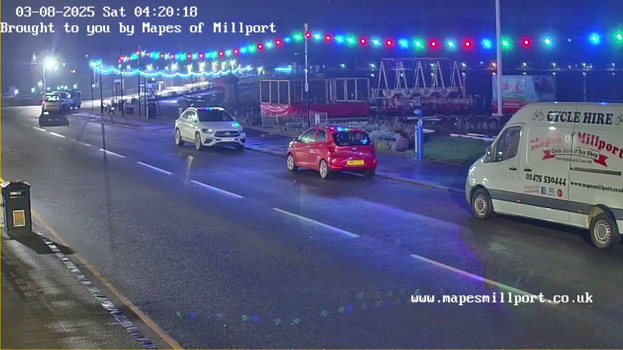 At night, three cars and a van are parked or driving on a wet road lined with colorful string lights.