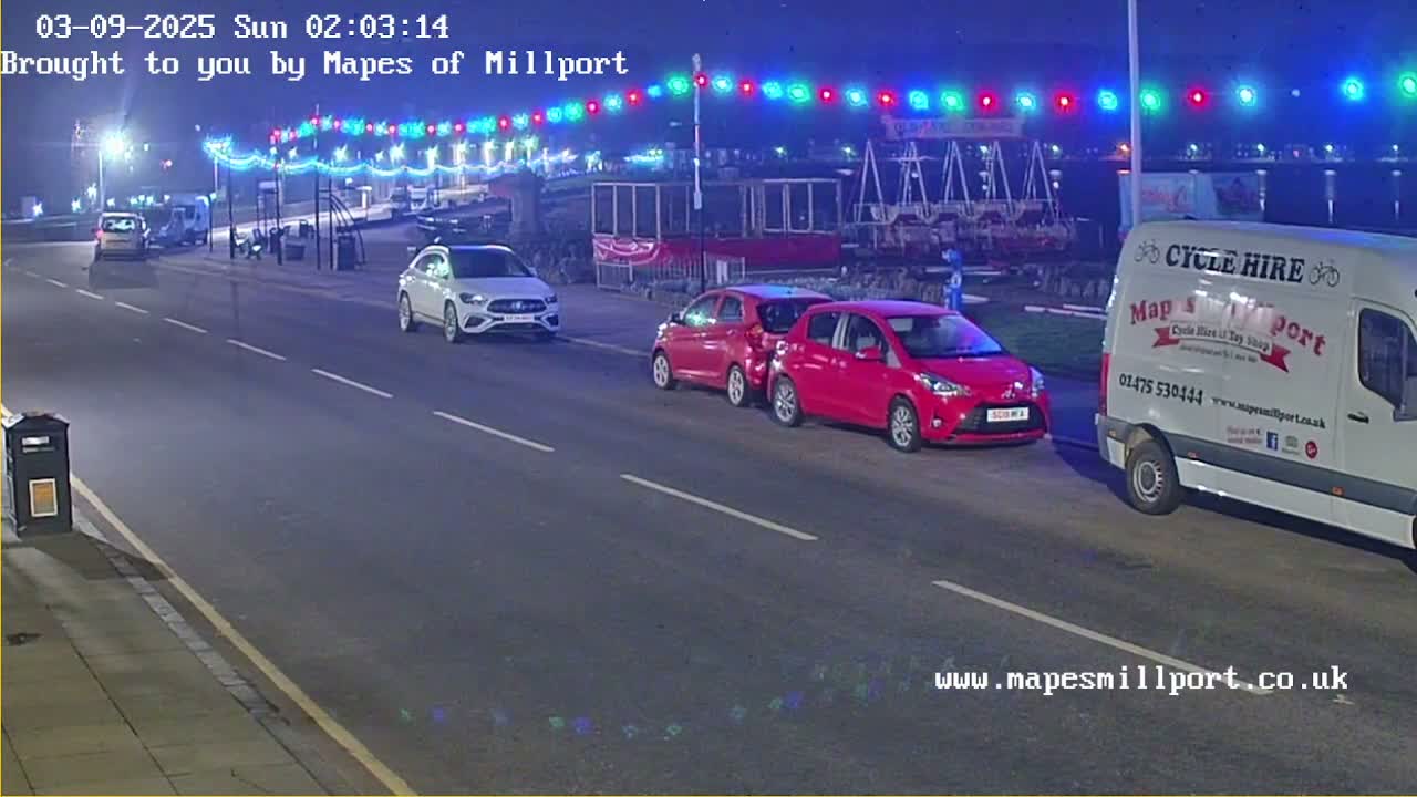 At night, several cars are parked along a road illuminated by multicolored lights strung overhead.
