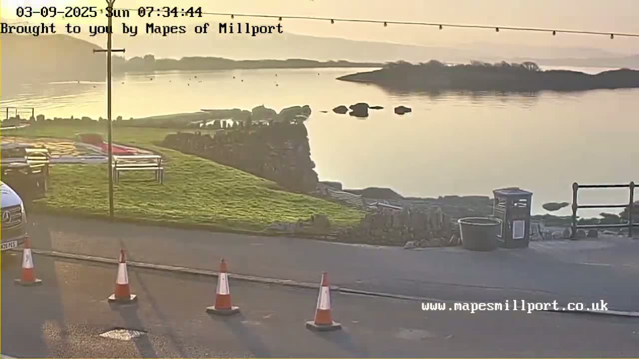 A calm, sunlit body of water is seen from a coastal vantage point, with grassy areas, a road, and traffic cones in the foreground.