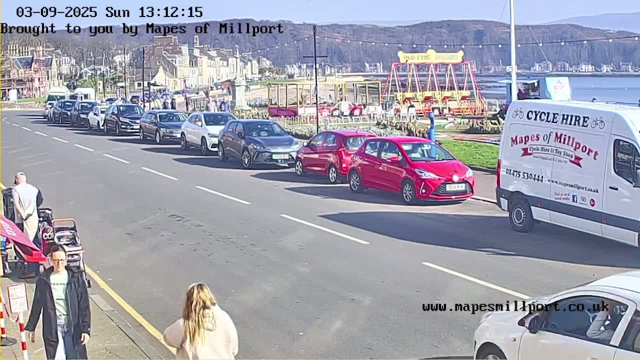 A sunny seaside street scene shows a line of parked cars, pedestrians, and a cycle hire van near a fairground attraction.