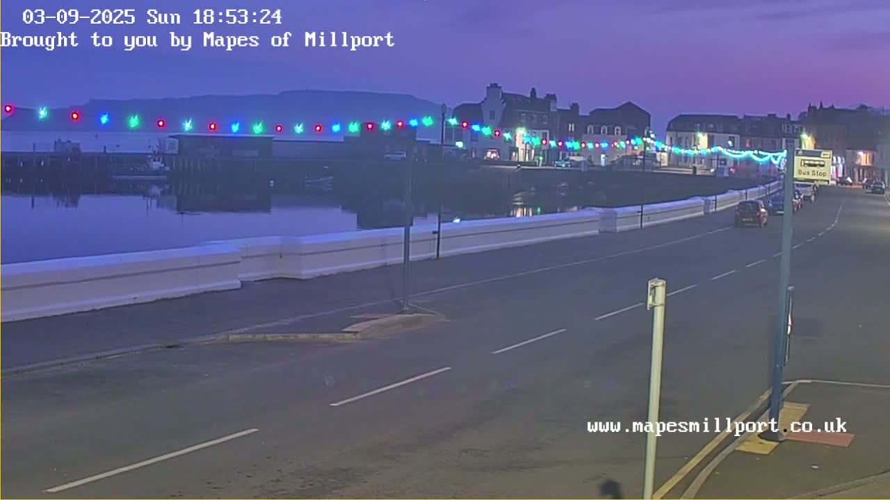 A twilight scene shows a waterfront road with multicolored lights strung along a low wall overlooking calm water, with cars on the road and buildings in the background.