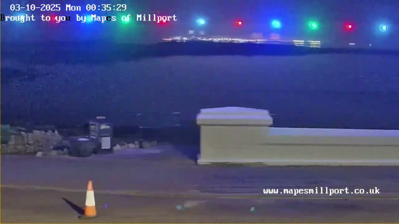 A nighttime view of a dark body of water with city lights in the distance, a low wall, and an orange traffic cone in the foreground.