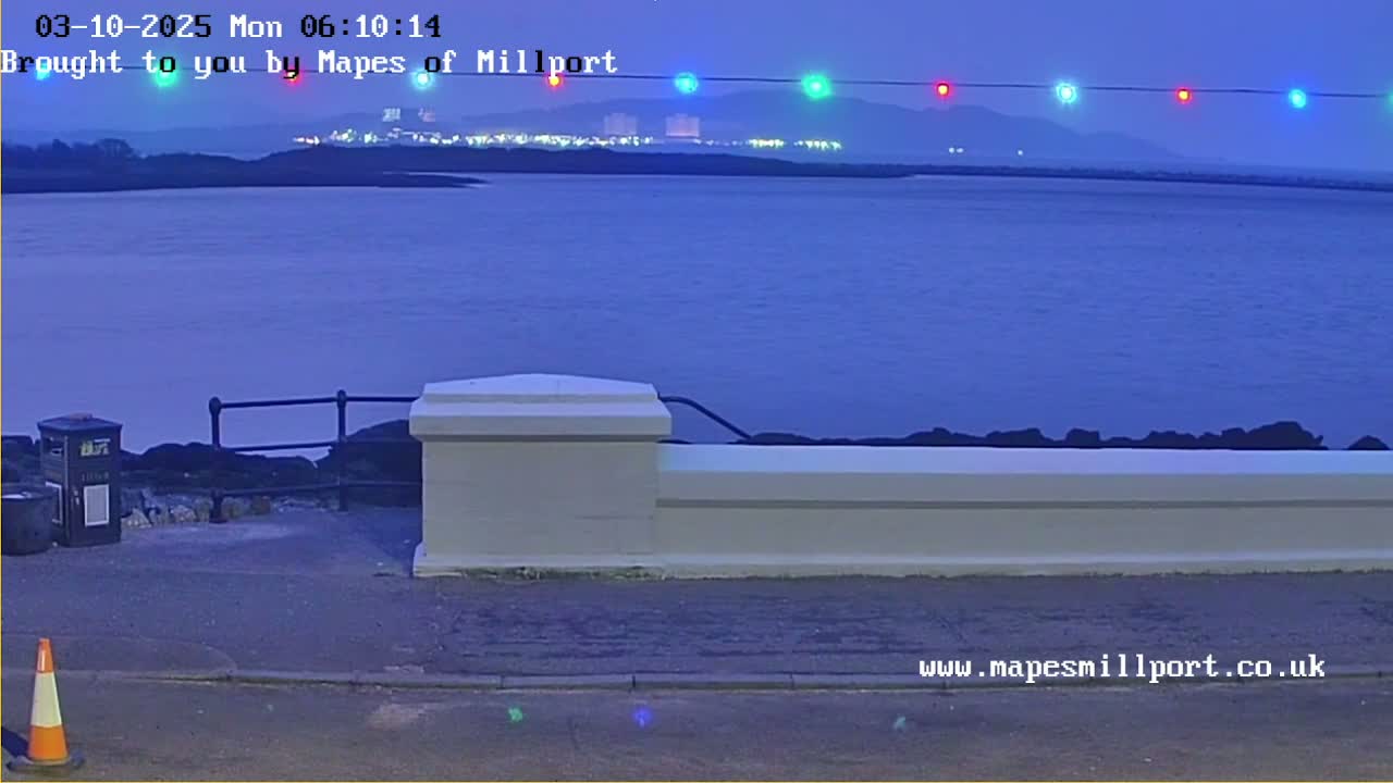 A twilight view of a calm body of water with a low white wall and a distant illuminated shoreline.