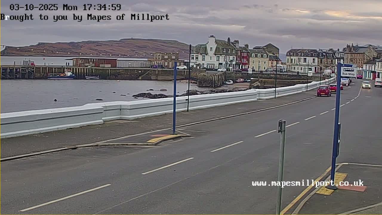 A road runs alongside a harbor, with several cars driving, and a low, white wall separating the road from the water; overcast skies are visible.