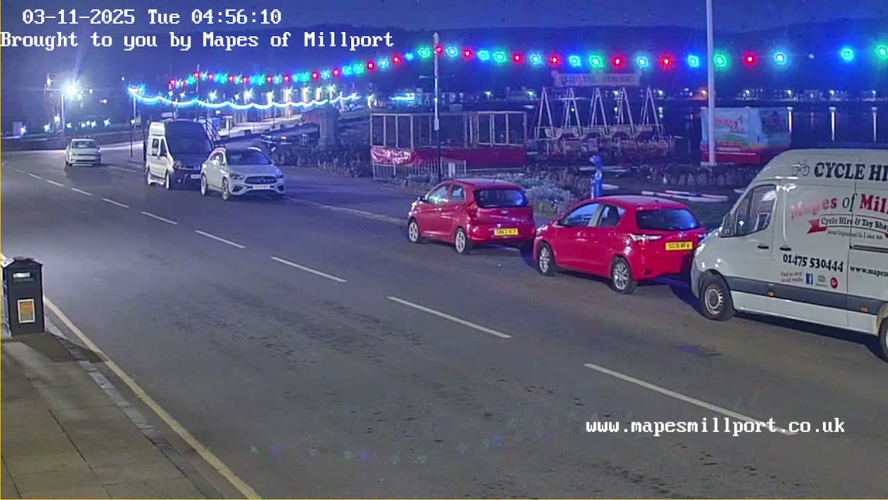 At night, several cars and vans are parked along a road lined with colorful lights, under a dark sky.