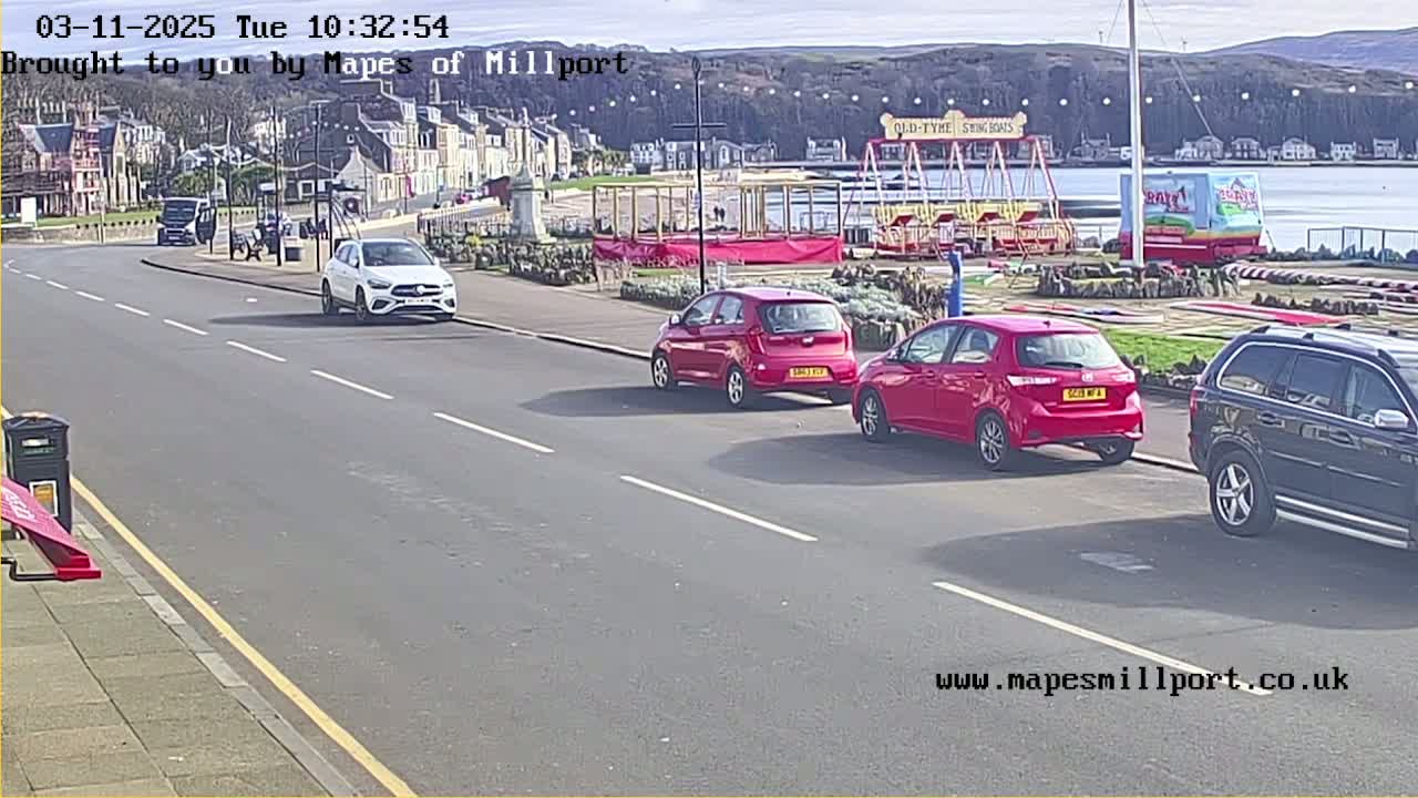 A sunny seaside street scene shows several cars parked and driving near a carousel and other fairground attractions.