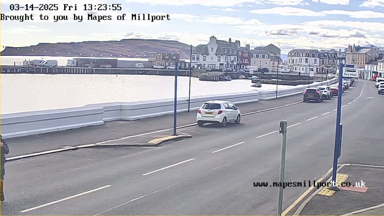 A seaside town with calm water, several cars parked along the road, and a few cars driving, under a mostly clear sky.