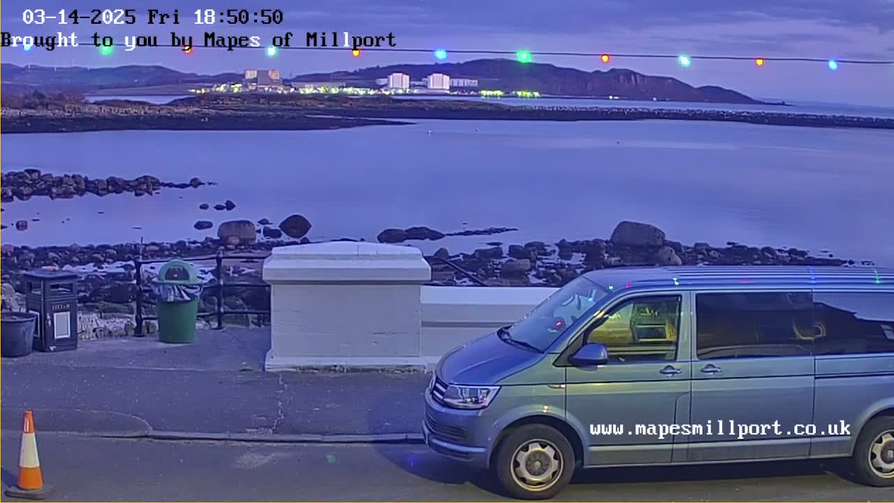 A light-blue minivan is parked near a rocky shoreline at dusk, overlooking a body of water and a distant town under a twilight sky.