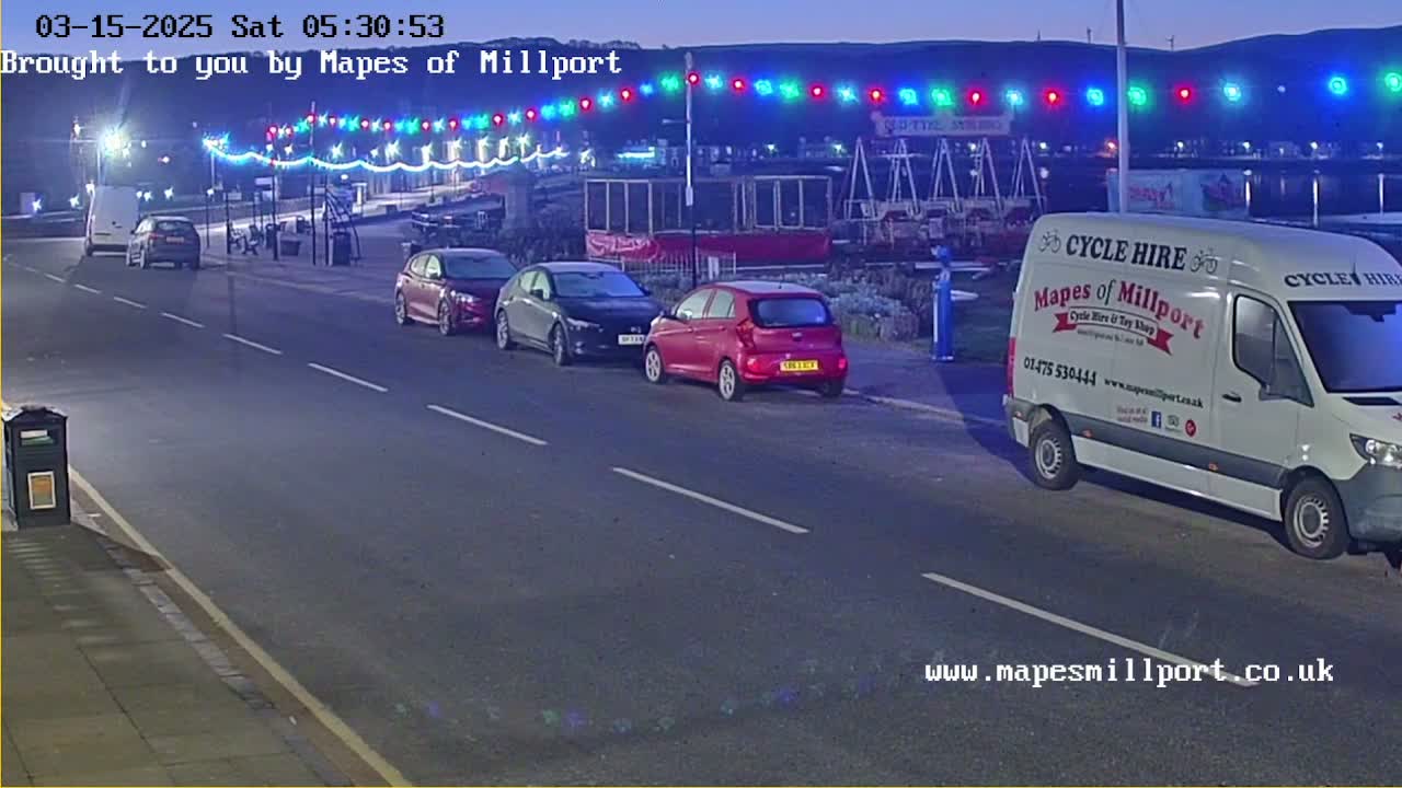 A mostly empty road at night with several parked cars and a van, under a string of multicolored lights, in twilight conditions.
