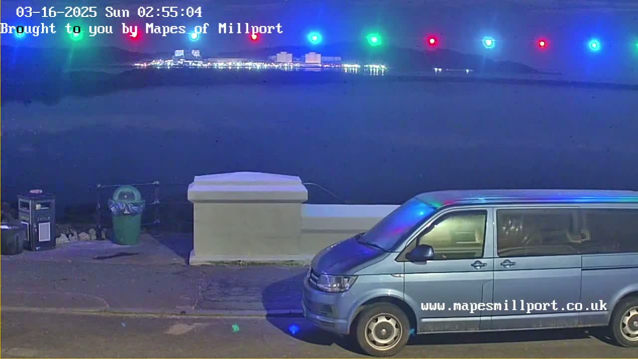 A light-blue van is parked near a low wall overlooking a dark body of water at night, with a distant town illuminated.
