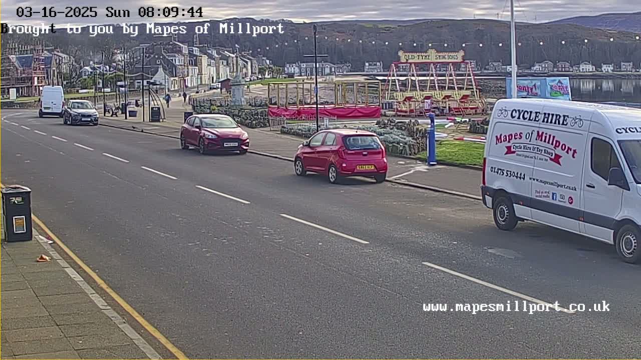 A street scene shows two red cars and a white van parked near a seaside town with an overcast sky, a carousel, and a few pedestrians visible in the distance.