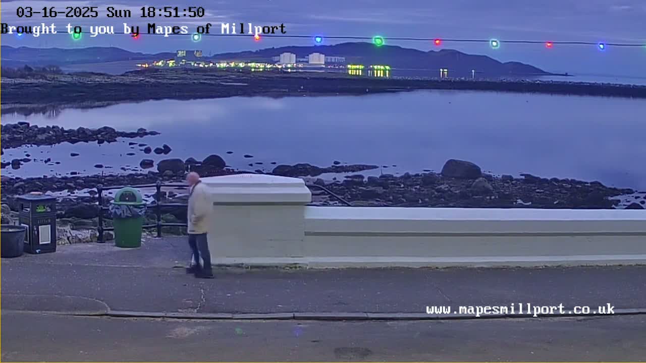 A man in a light-colored coat stands near a low wall overlooking a calm body of water at dusk, with a rocky shoreline and distant lights visible.