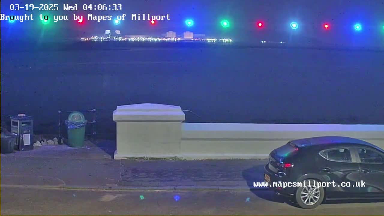 A dark-colored car is parked near a low wall overlooking a dark body of water at night, with city lights visible in the distance.