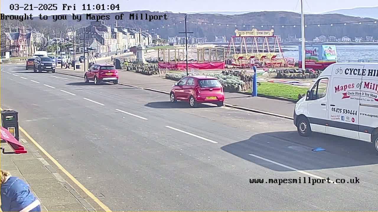Several cars are parked along a seaside road on a sunny day, near a playground and a van.
