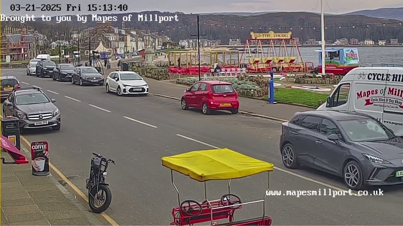 A street scene shows several cars parked and driving along a road near a waterfront with a carousel and other fairground rides under an overcast sky.