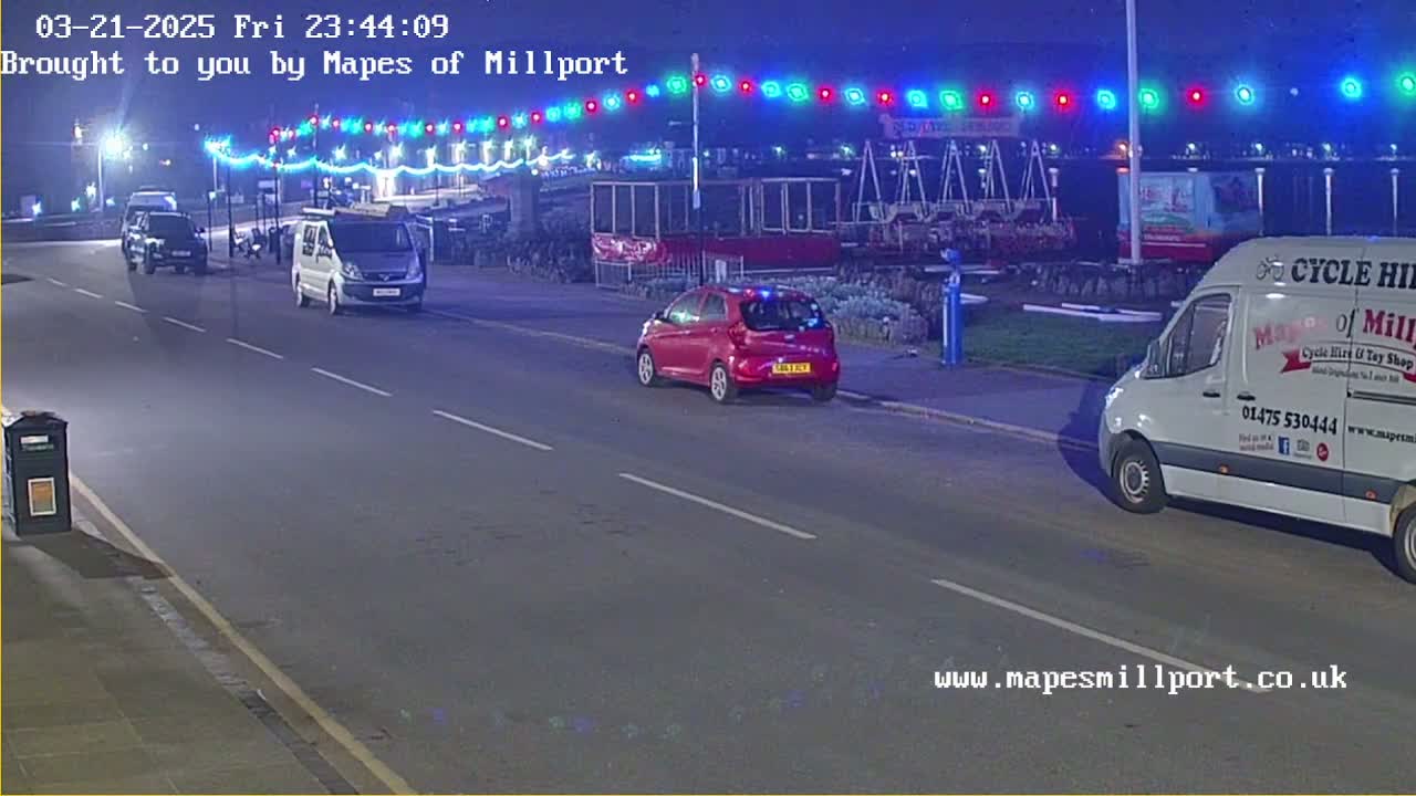 At night, under multicolored string lights, several vehicles are parked and driving on a road near a seaside amusement area.