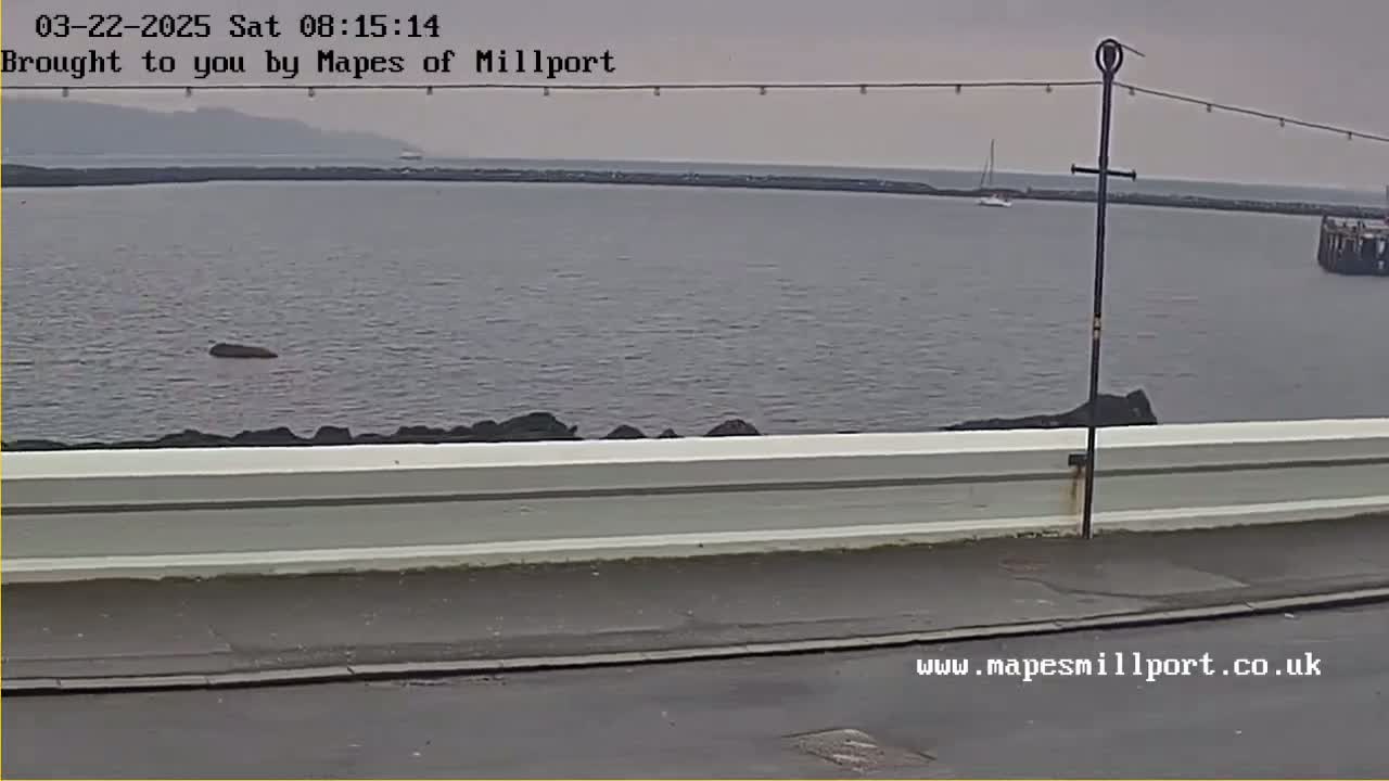 A calm, gray day is shown overlooking a body of water with a low concrete wall and a single sailboat in the distance.
