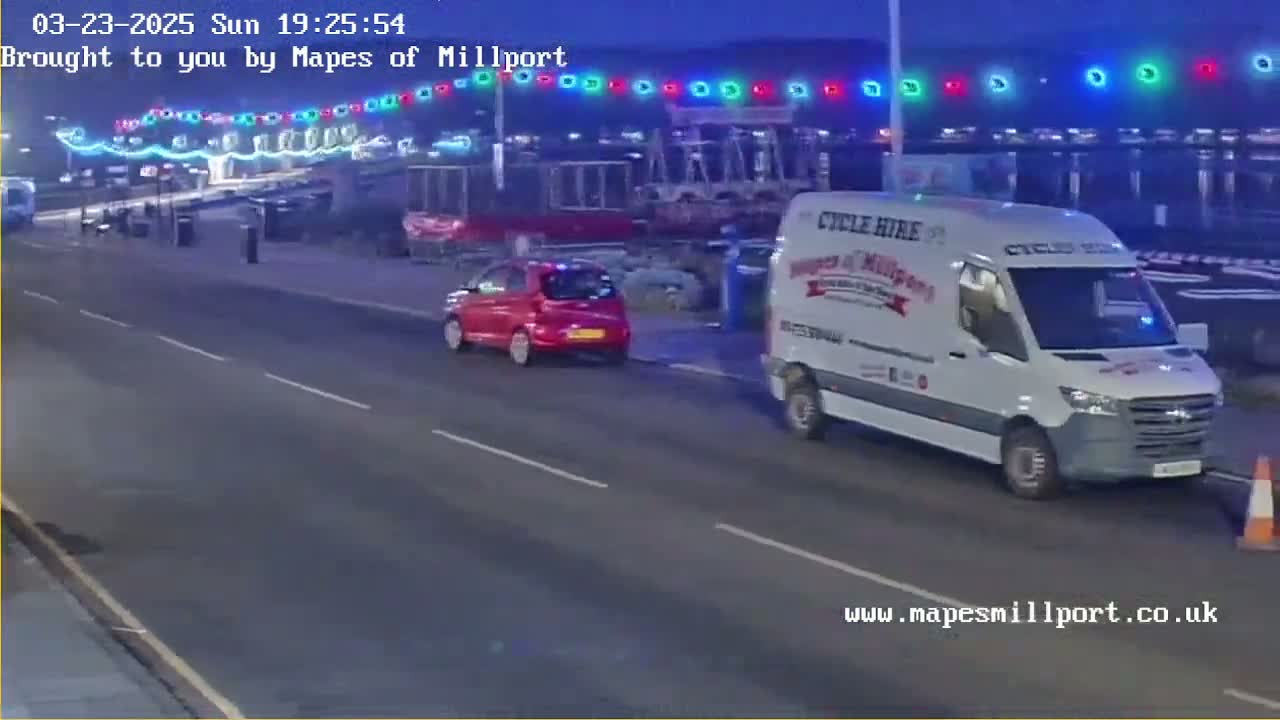 A red car and a white van are driving on a road at night, under a string of colorful lights.