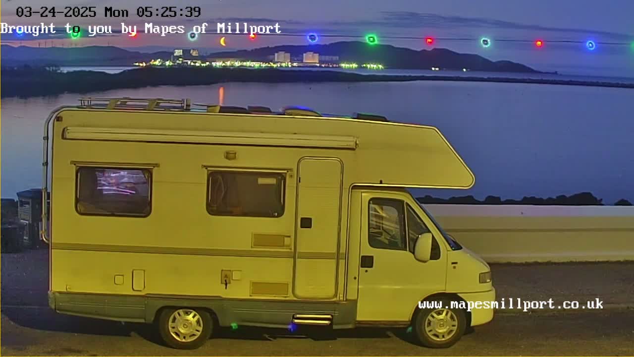 A yellowish campervan is parked near a body of water at twilight, with a string of colored lights visible in the distance across the water.