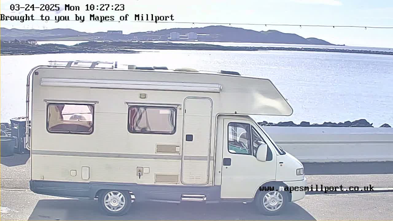 A white campervan is parked near a body of water on a sunny day with a distant shoreline visible.