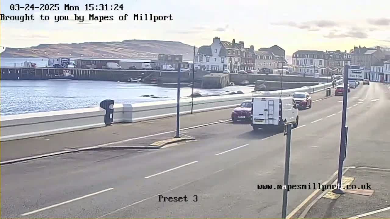 A sunny day reveals a harbor scene with several cars driving along a road that runs parallel to a waterfront, where boats are docked near buildings.