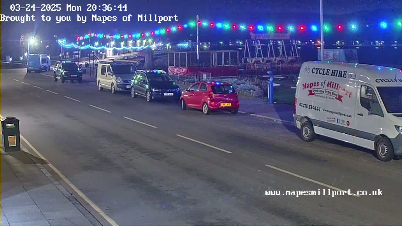 Several cars are parked along a road at night under a string of colorful lights.