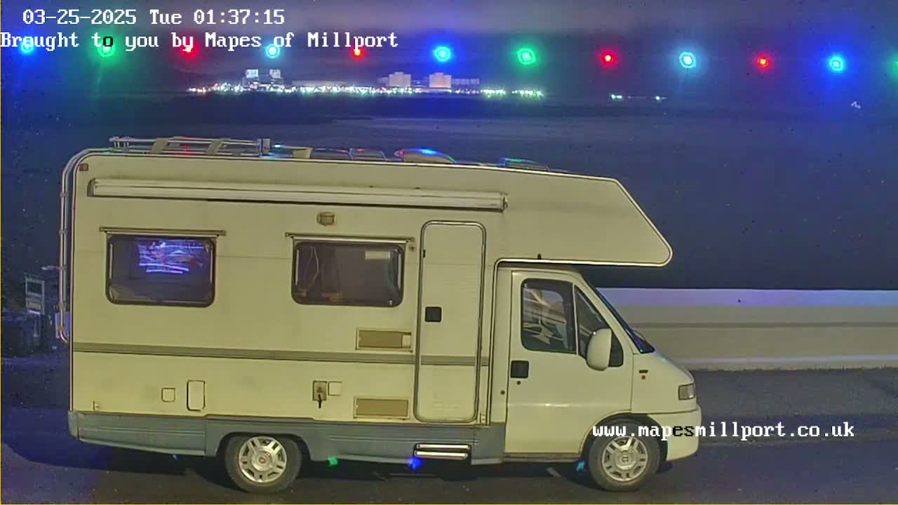 A white campervan is parked at night near a body of water with a distant line of lights visible.