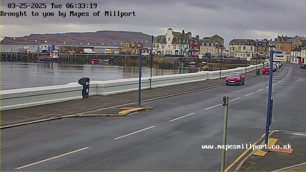 A harbor town under an overcast sky is seen from across the street, with several cars parked and driving along a road beside a low white wall.