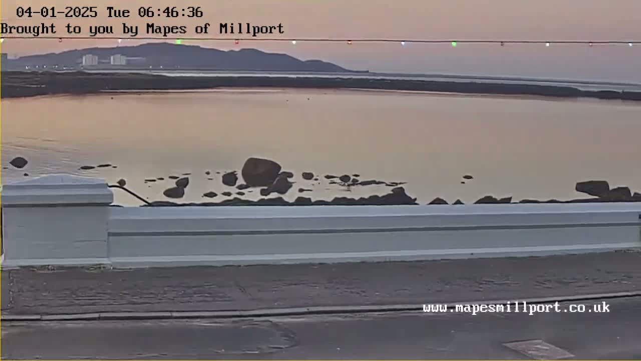 A calm body of water, appearing pinkish from the sunset, stretches out before a low white wall, with dark rocks visible in the water near the shore.
