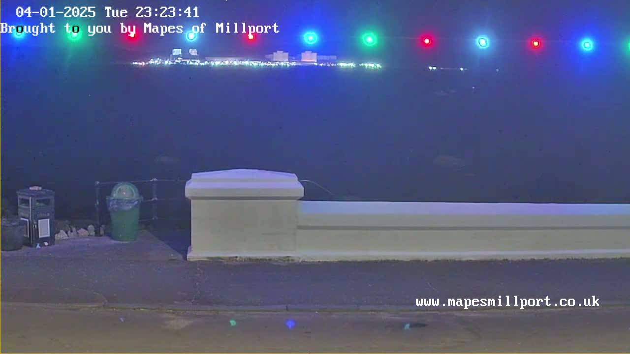 A hazy, nighttime view of a waterfront, showing a low wall, trash receptacles, and distant lights across the water.