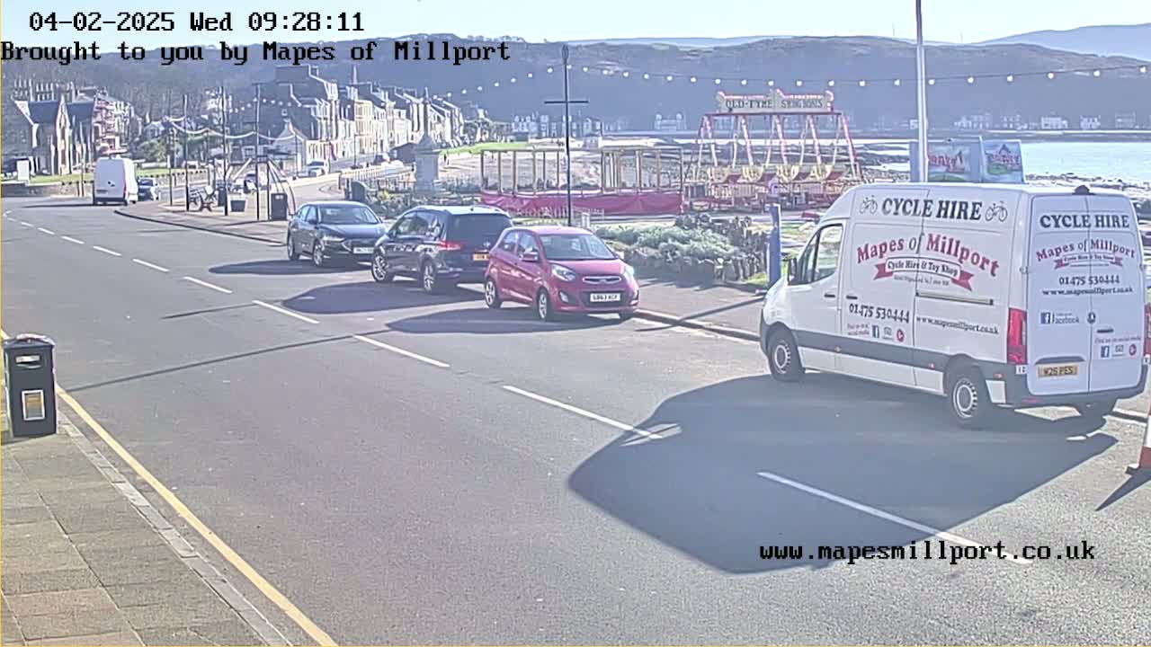 A sunny seaside street scene shows several parked cars, a cycle hire van, and a carousel in the background.