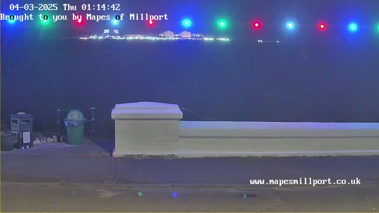 A nighttime view of a body of water with lights across the far shore, seen from behind a low white wall with trash receptacles nearby under hazy conditions.