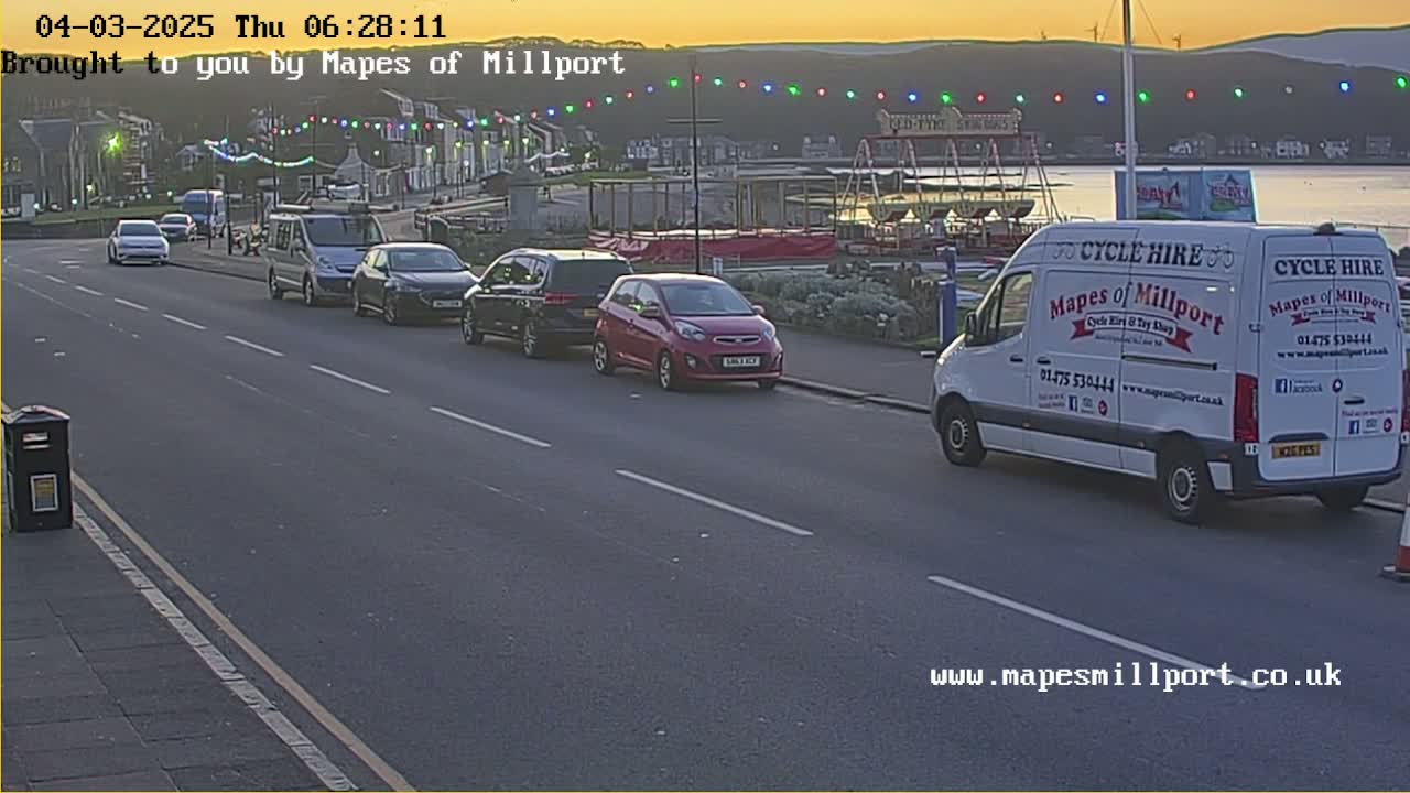 Several cars and a van are parked along a seaside road at sunset, with a fairground ride visible in the background.