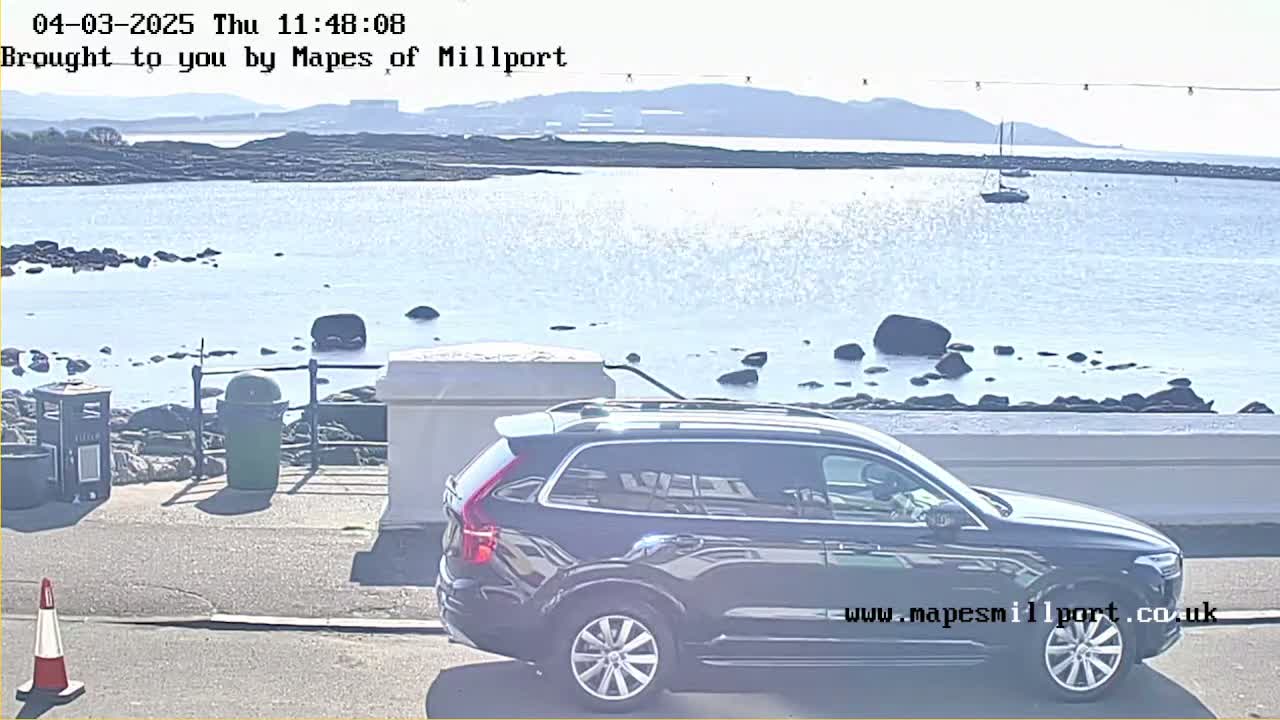 A dark-colored SUV is parked near a waterfront with calm water, rocks, and distant mountains under a sunny sky.