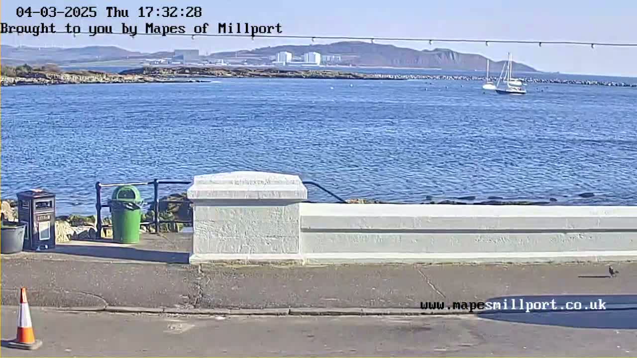 A sunny day shows a calm body of water with a sailboat, a low white wall, and trash cans along a paved area.