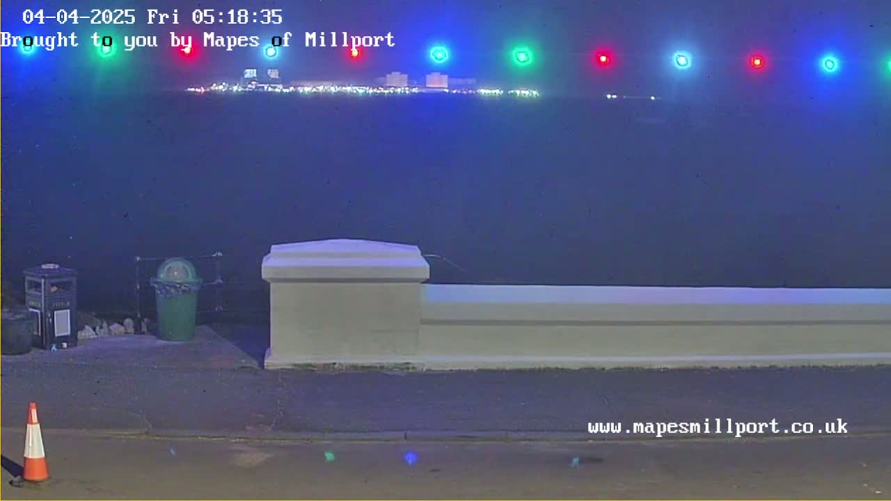 A nighttime view shows a body of water with faintly lit buildings in the distance, a low white wall in the foreground, and a traffic cone and two trash cans near the wall under a dark, hazy sky.