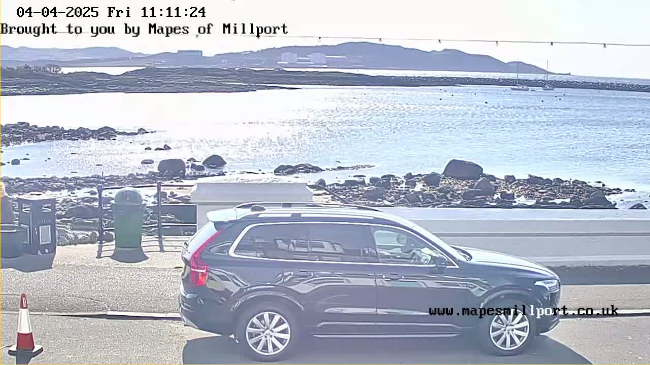 A dark-colored SUV is parked near a rocky shoreline overlooking a calm body of water on a sunny day.