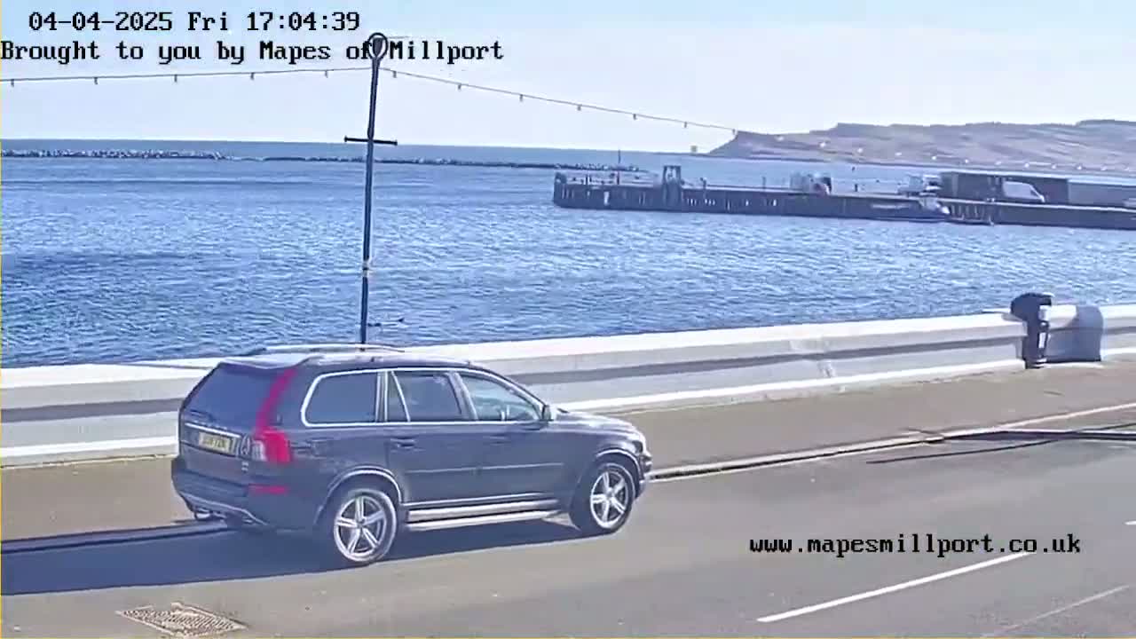 A dark-colored SUV drives along a road next to a body of water on a sunny day.