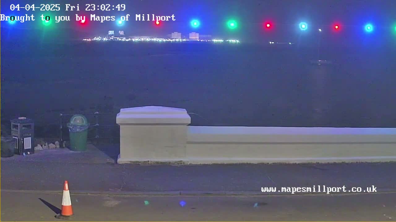 A nighttime view of a waterfront scene with hazy conditions, featuring a low wall, trash receptacles, a traffic cone, and distant lights across the water.