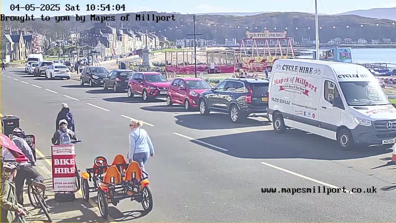 A sunny street scene shows several cars parked and driving, a few pedestrians, a bike hire van, and a person pushing a four-wheeled orange bike.