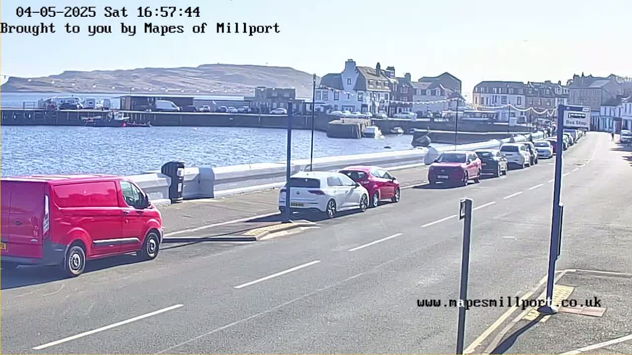 A sunny day reveals a red van parked near a waterfront road lined with cars, overlooking a harbor with buildings in the background.