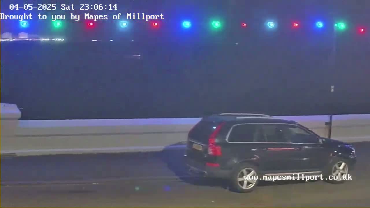 A dark-colored SUV is parked on a road at night in foggy conditions, with a row of multicolored lights in the background.