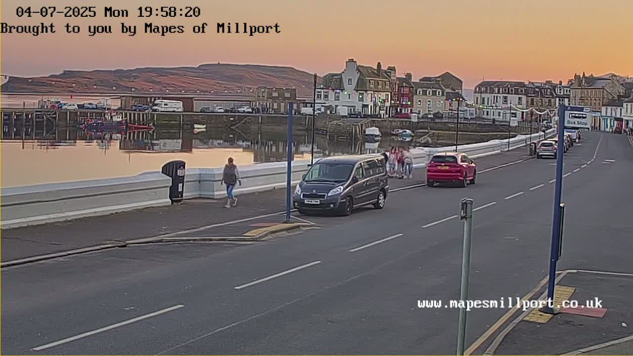 A seaside town at sunset, with several people walking along a waterfront road beside a calm body of water, where a dark-colored minivan and a red car are parked.