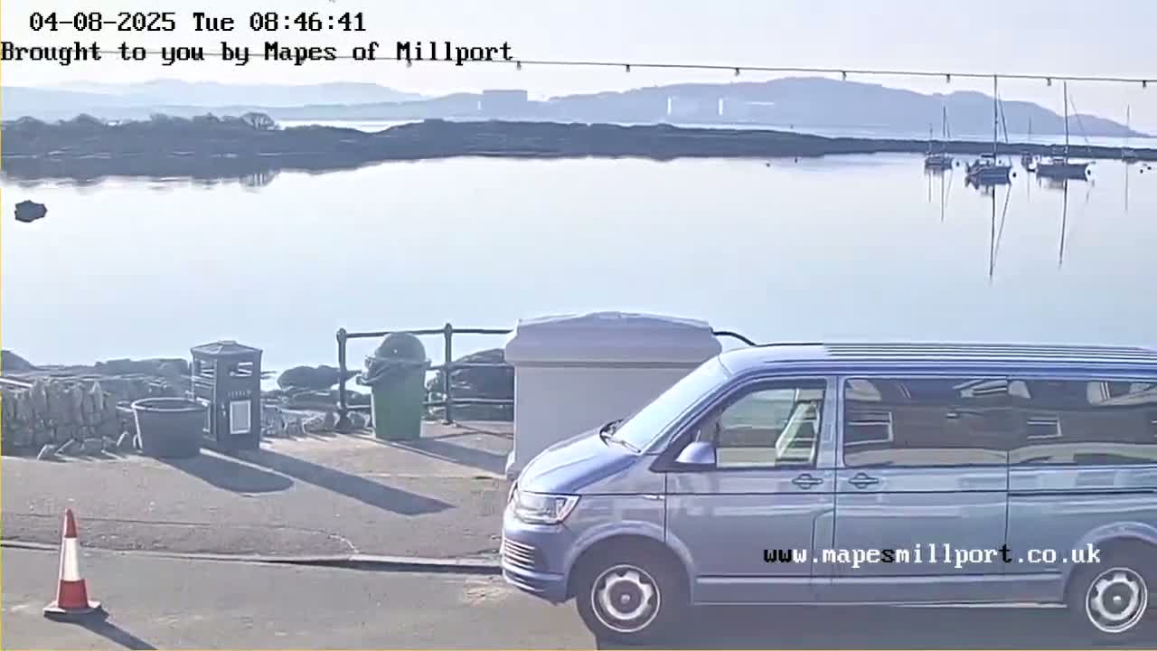 A silver minivan is parked near a waterfront on a sunny day with several boats visible in the calm water and distant hills in the background.