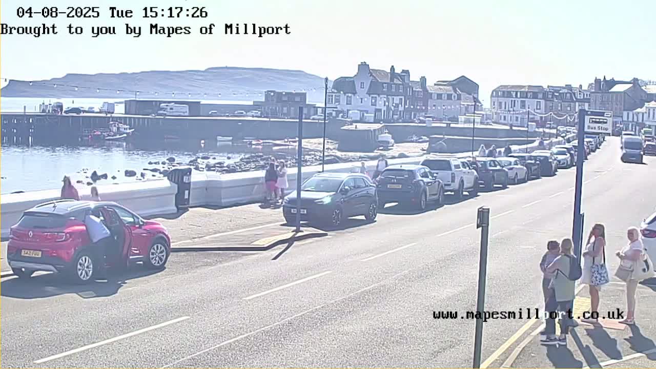 A sunny day shows a seaside town with parked cars lining a road near a waterfront, where several people are walking near a low wall.