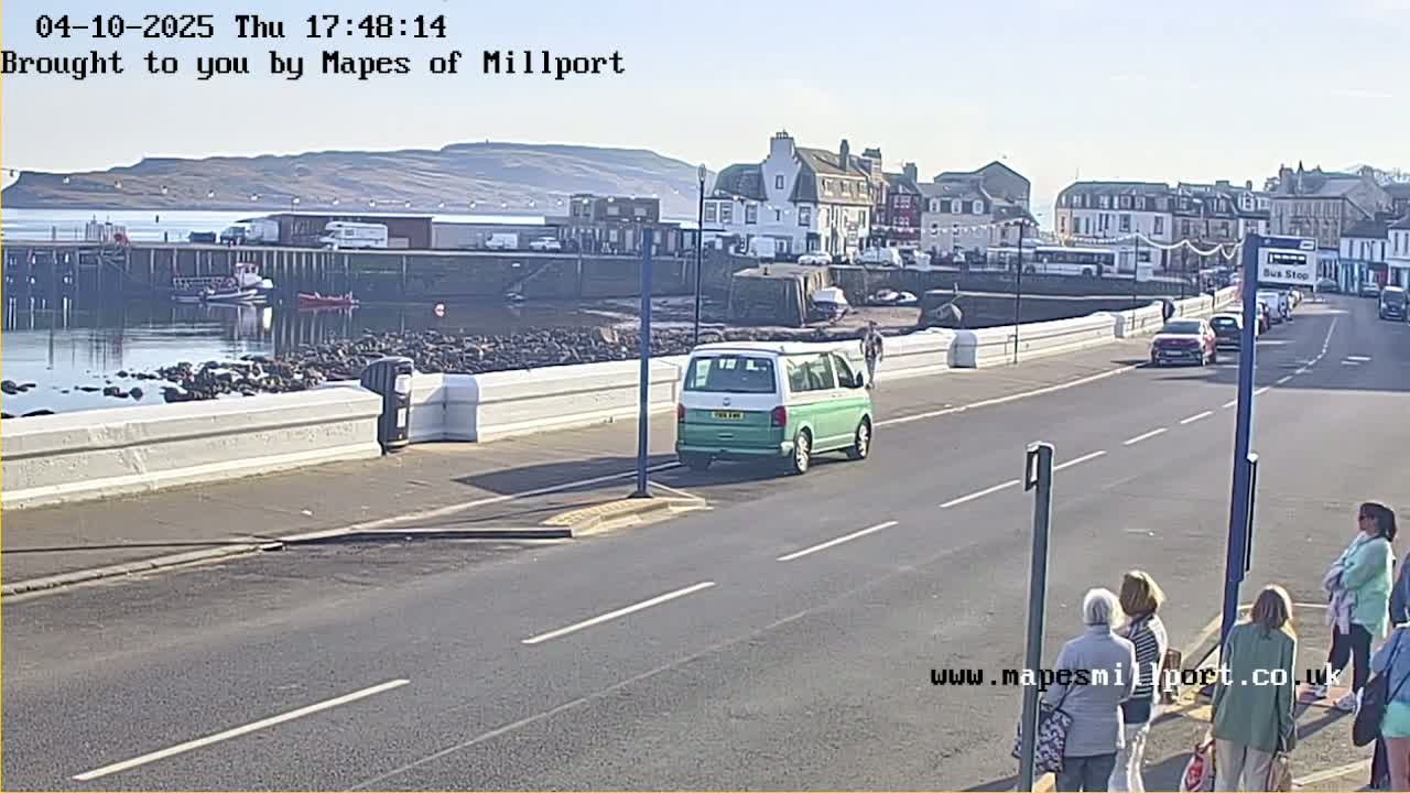 A light-green van is driving on a road next to a harbor on a sunny day, with several people standing at a bus stop and boats in the water.