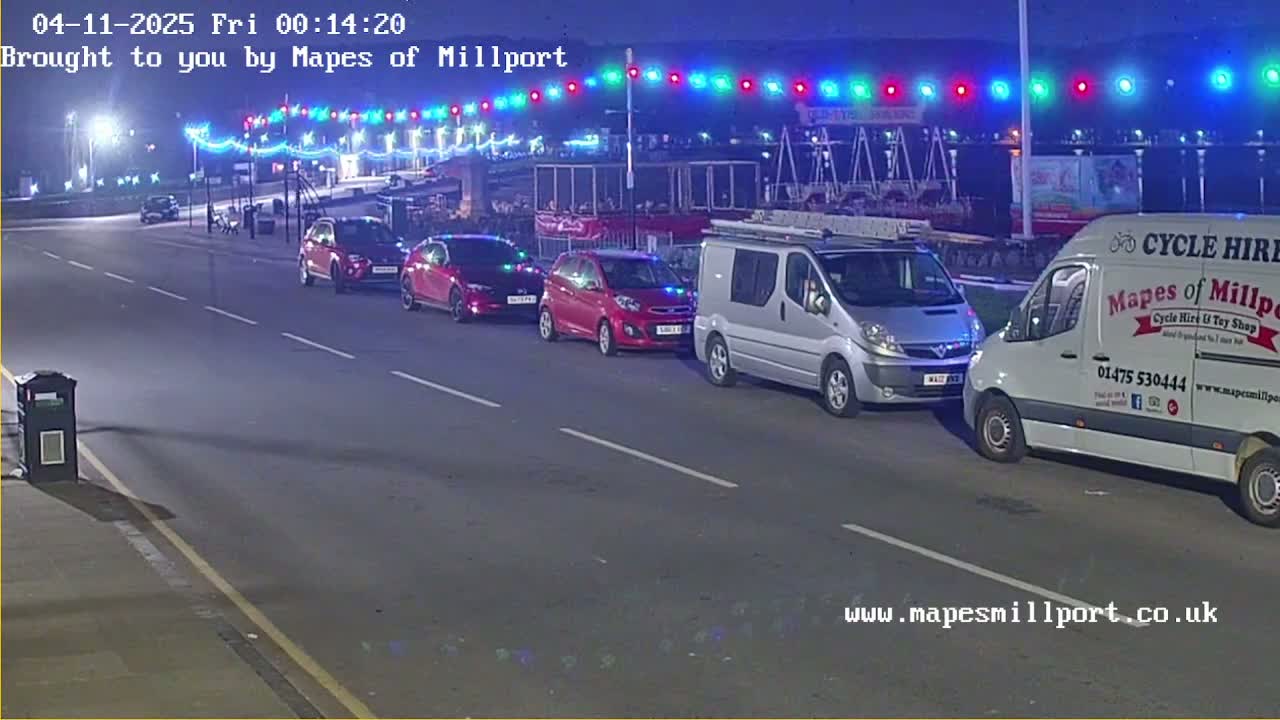 Several cars and vans are parked along a road at night under a string of colorful lights.