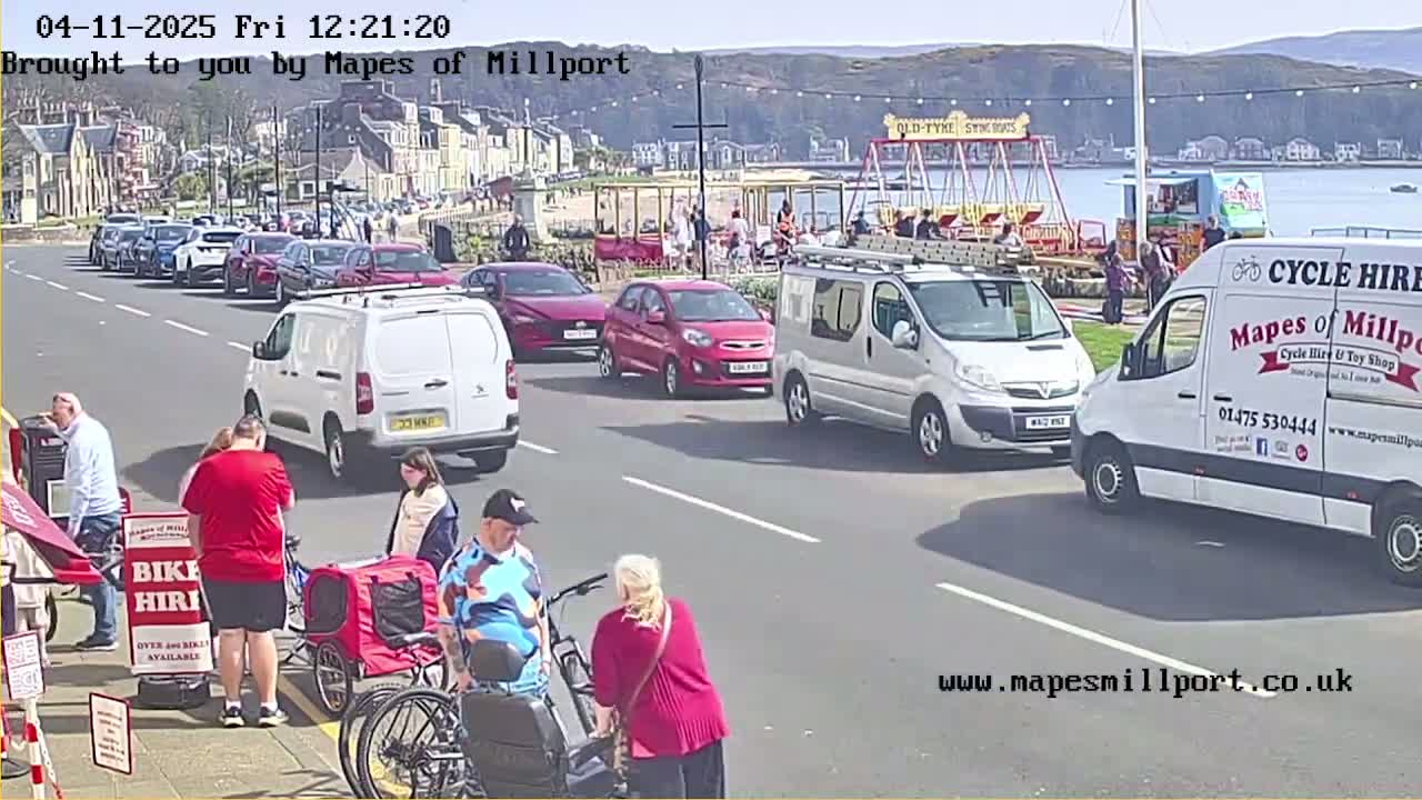 A seaside street scene on a sunny day shows parked cars, pedestrians, and several vans, including one advertising cycle hire, near a playground and body of water.