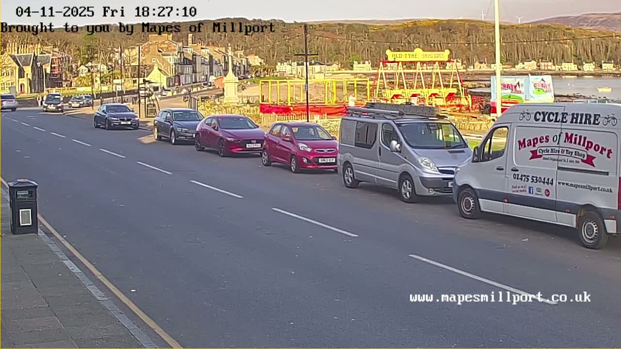 Several cars are parked on the side of a road in front of a seaside town under a clear, bright sky.