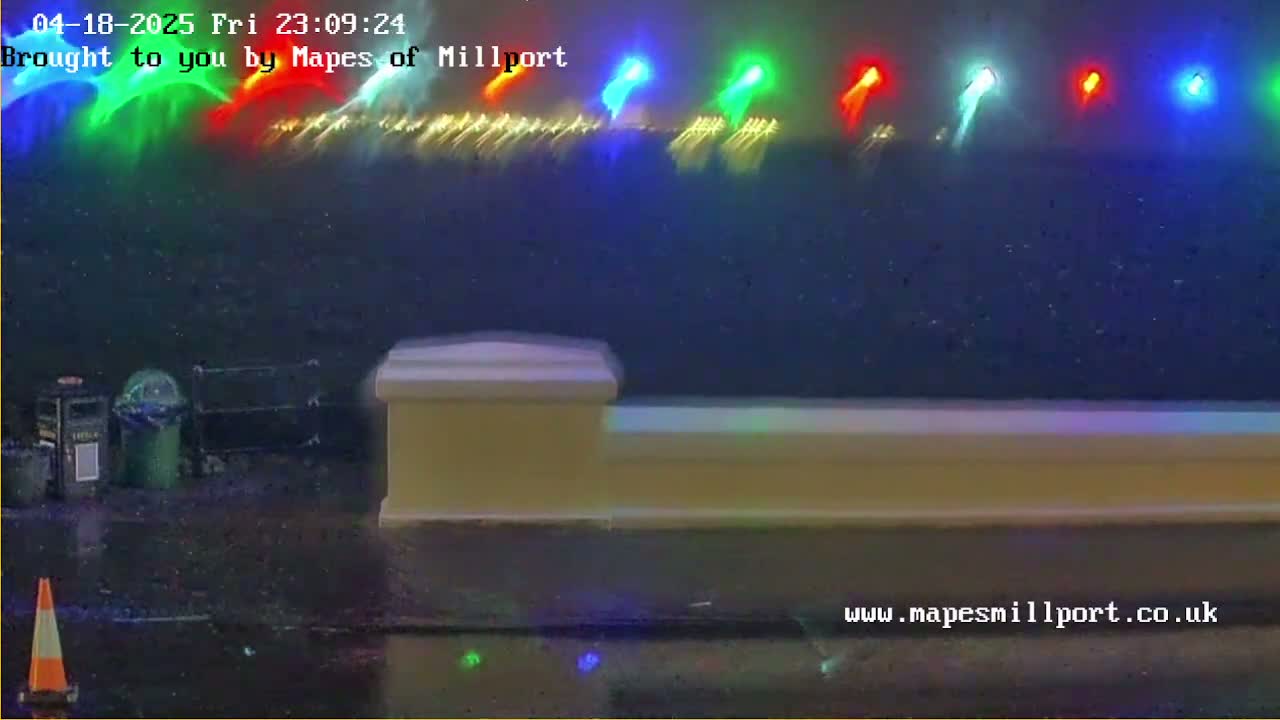 A low-light, nighttime outdoor scene shows a low, tan-colored wall with multicolored lights blurred in the background, and a traffic cone in the foreground on a wet surface.